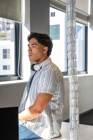 Asian man working on laptop in bright coworking space wearing headphones and striped shirt