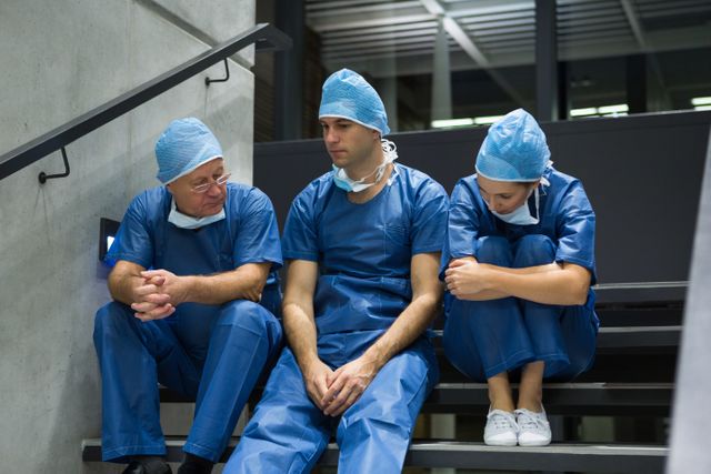 Group of surgeons in blue scrubs and surgical caps sitting on a staircase in a hospital, appearing worried and stressed. This image can be used to depict the emotional challenges and pressures faced by healthcare professionals, teamwork in medical settings, or the importance of emotional support among medical staff.
