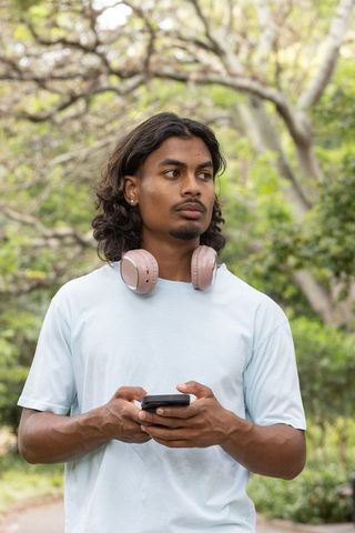 Young indian man with pink headphones holding smartphone in park, casual blue t-shirt