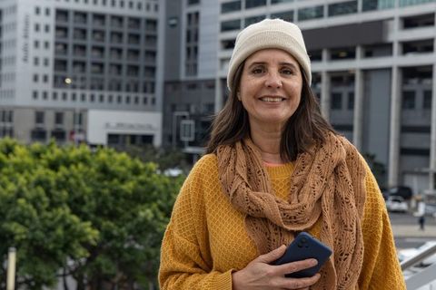 Smiling middle-aged woman on city terrace wearing beanie scarf and holding smartphone
