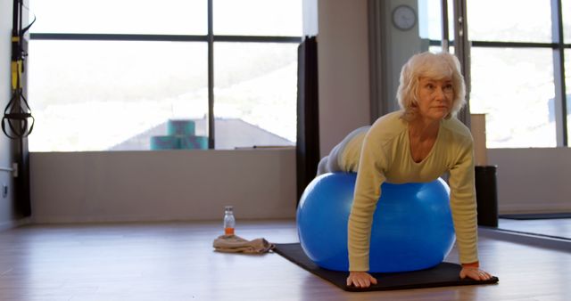 A senior Caucasian woman is exercising on a blue fitness ball in a bright room, with copy space. Her commitment to maintaining an active lifestyle is evident through her engagement in physical activity.