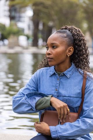 Contemplative african american woman at pond wearing denim shirt holding leather bag