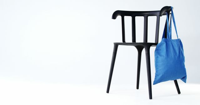 A black chair stands against a white background, with a blue tote bag hanging on its back, with copy space. Its simplicity suggests a minimalist design concept or the idea of bringing your own bag to reduce plastic use.