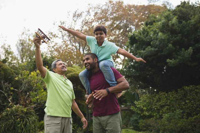 Three generations of a family enjoying time together in a park. The grandfather is holding a toy airplane, the father is carrying his son on his shoulders, and the son is pretending to fly. This image can be used for advertisements, family-oriented content, parenting blogs, and articles about family bonding and outdoor activities.