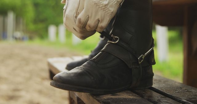 Person tightening riding boots strap, wearing gloves, in preparation for horse riding. Can be used for equestrian sport promotions, riding gear advertisements, horse riding tutorials, or outdoor activity themes.