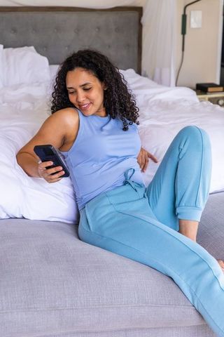 African american woman relaxing on bed edge in blue loungewear scrolling smartphone
