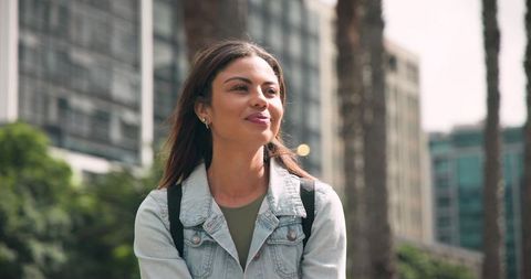 Confident young woman smiling and gazing in city park wearing denim jacket and backpack