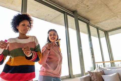 Friends cleaning glass doors in sunlit modern living room, dancing and laughing together