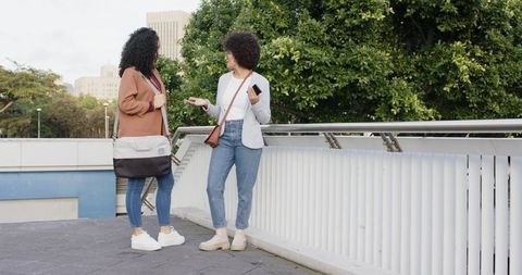 African american women chatting on pedestrian bridge carrying messenger bag and smartphone