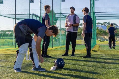 Young cricketer adjusting pads at sunlit practice nets with coach and teammates