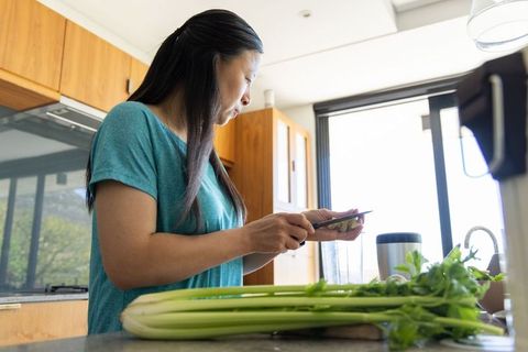 Asian woman preparing celery for green smoothie in sunlit modern kitchen