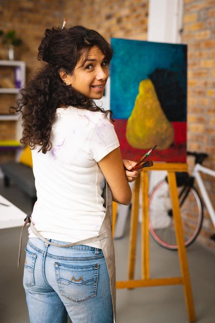 Portrait of happy biracial female artist with painting equipment and apron in creative office. Female artist studio and painting concept.