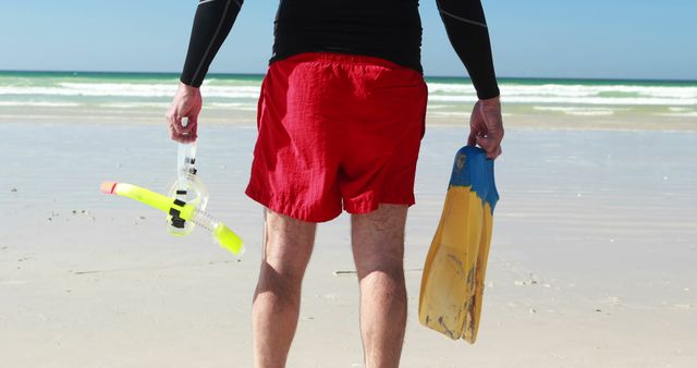 Man standing on sandy beach, holding snorkeling mask and fins, looking towards ocean. Ideal for use in travel brochures, beach holiday promotions, adventure sports advertisements, and summer vacation-themed content.