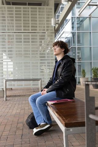 Teenager wearing leather jacket sitting on bench in glass atrium holding notebooks urban plaza