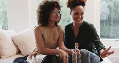 Middle-aged African American women sharing drinks and laughter on cozy living room sofa