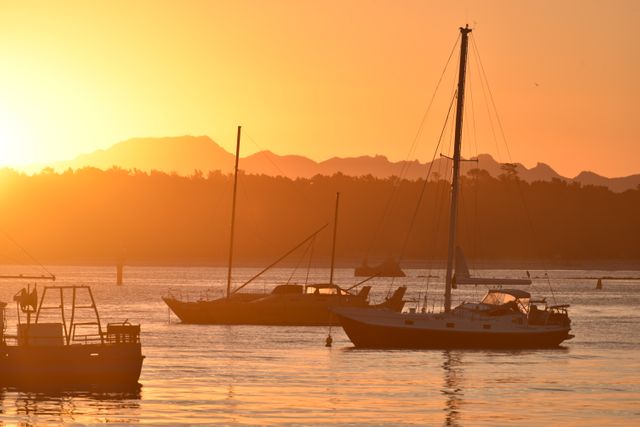 Sailboats resting on calm water as the sun sets, casting a golden glow over the scene. Mountains and trees in the background. Suitable for use in travel promotions, nature retreats, or lifestyle blogs.