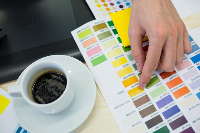 Hands of a male graphic designer selecting colors from a color chart in an office. A cup of coffee is placed nearby, indicating a focused and creative work environment. Ideal for use in articles or advertisements related to graphic design, creative processes, office work, and professional design services.