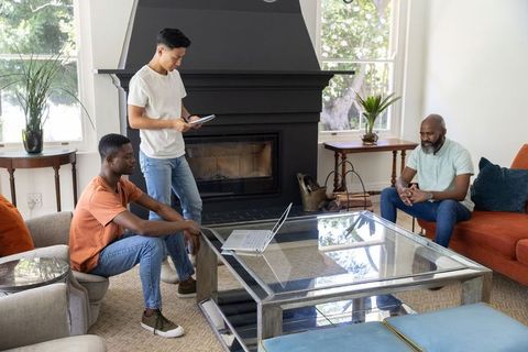 Multiracial men discussing project in cozy living room around glass coffee table with laptop