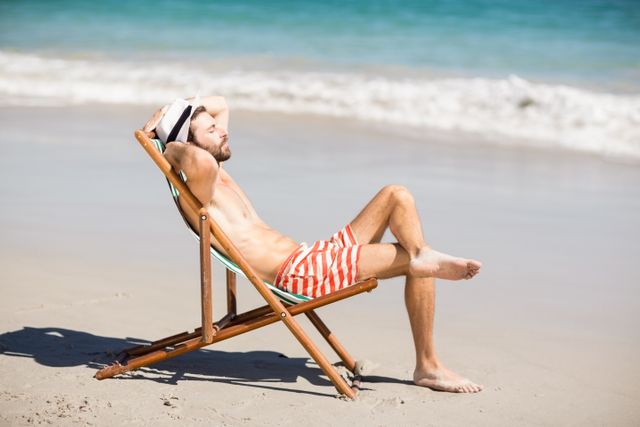 Young man enjoying a sunny day at the beach, sitting on a wooden beach chair near the ocean. Ideal for use in travel brochures, vacation advertisements, lifestyle blogs, and summer-themed promotions.
