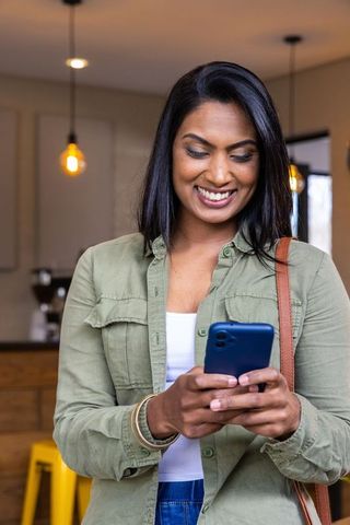 Indian woman smiling while scrolling smartphone in cozy coffeehouse with warm lighting