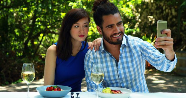 A young Caucasian couple enjoys a meal outdoors and takes a selfie, with copy space. Capturing memories, they share a moment of togetherness during a romantic lunch.