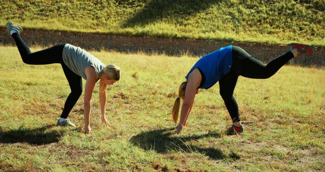 Two young Caucasian women are engaged in outdoor fitness, performing exercises on a grassy field, with copy space. Their athletic attire and synchronized movements suggest a focus on health and physical activity.