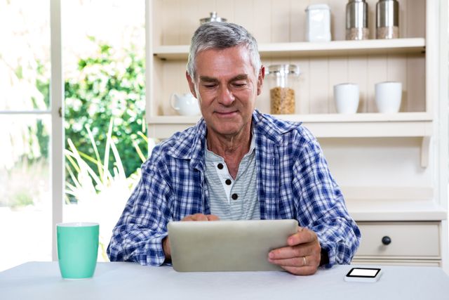Senior man sitting in kitchen using digital tablet. Ideal for illustrating concepts of technology use among elderly, modern lifestyle, home activities, and digital communication. Can be used in articles, advertisements, and websites focused on senior living, technology adoption, and home life.