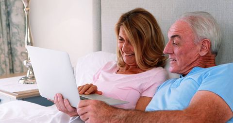 Happy Senior Couple Relaxing in Bed with Laptop in Bedroom