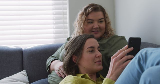 Caucasian lesbian couple using smartphone and embracing on couch. domestic life, spending free time relaxing at home.