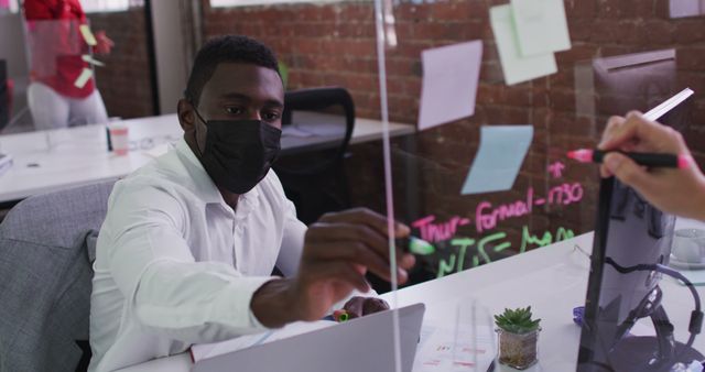 Businessman wearing black mask writing on transparent board in office. Suitable for visuals emphasizing safe workplace practices, team collaboration during pandemic, and productivity in modern office environments.