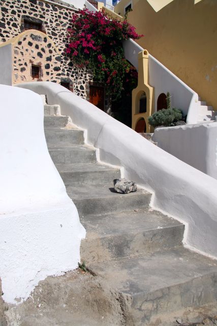 Rustic stone stairway leading to traditional Greek house with vibrant bougainvillea and distinctive white walls. Suitable for travel brochures, cultural heritage sites, Mediterranean lifestyle blogs, or vacation planning materials highlighting Greek architecture and scenic beauty.