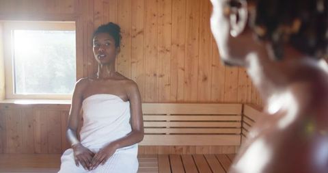 African american couple relaxing in sunlit wooden sauna, woman wearing towel
