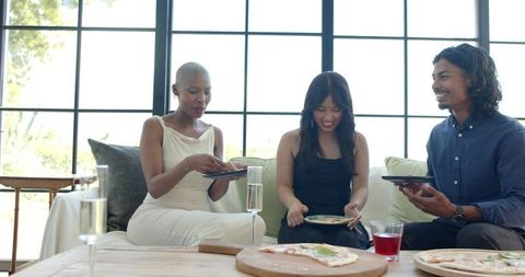 Friends sharing pizza on sunlit sofa, casual home gathering, laughing multicultural group