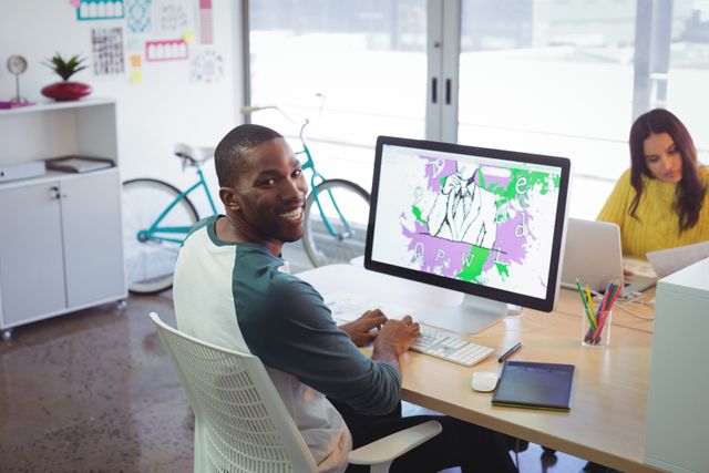 Male designer smiling while working on computer in a modern creative office. Ideal for use in articles about creative workspaces, teamwork, graphic design, and modern office environments.