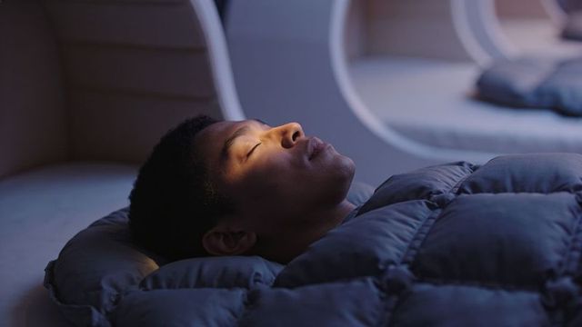 Young man resting in sleep capsule under dark quilted comforter, moody ambient lighting