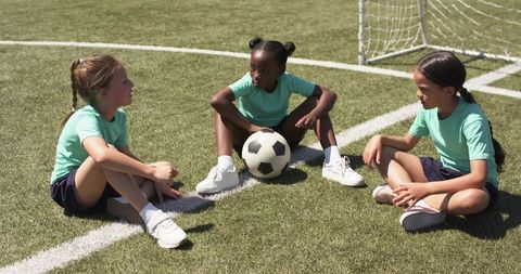 Youth girls sitting on soccer field holding ball during team talk, diverse junior football players