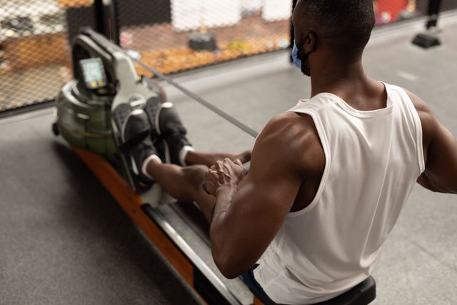 African American man exercising on a rowing machine in a gym while wearing a facemask and a white tank top. Suitable for illustrating fitness routines, health and safety measures in gyms, and home workout guides.