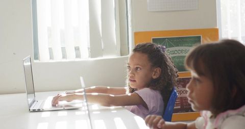 Sunlit classroom african american girl reaching for laptop keyboard while peer focusing