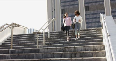Two women walking down city stairs carrying gym duffel bags and coffee during morning commute