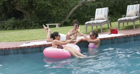 Friends toasting cocktails while relaxing on pink donut float in backyard pool