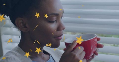 Woman holding red mug near window blinds with gold star overlays, cozy mindful moment