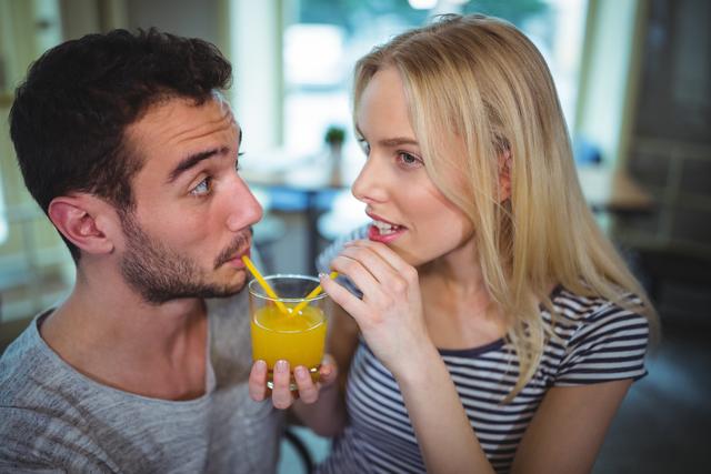 Young couple sharing orange juice in a cafÃ©, both smiling and enjoying their time together. This image can be used for themes related to relationships, romance, dating, refreshment, and leisure activities. Great for blogs, advertisements, social media, websites, and promotional materials centered around couples, beverages, and casual outings.