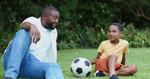 African american father and son sitting on lawn talking and playing with soccer ball