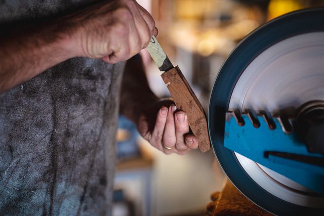 This image captures the hands of a Caucasian male knife maker using an angle grinder in a workshop. It is ideal for illustrating concepts related to craftsmanship, woodworking, metalworking, and small business. Suitable for use in articles, blogs, and advertisements focused on handmade products, DIY projects, and skilled trades.