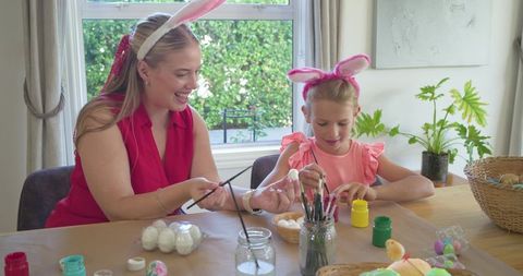 Mother and daughter painting easter eggs together at dining table wearing bunny ears