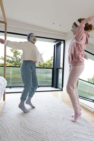 Friends jumping on rug wearing facial masks and cozy loungewear in sunlit modern bedroom