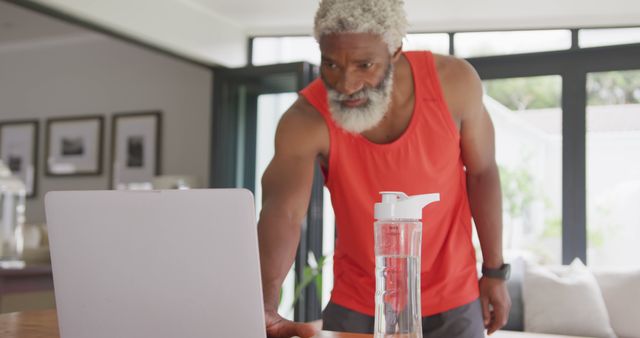 Senior man wearing a red tank top using a laptop in a modern living room. He appears engaged with a screen, likely following an online fitness program. A water bottle is on the table. This image can be used to represent healthy lifestyles, senior fitness, technology in exercise, and home workout scenarios.