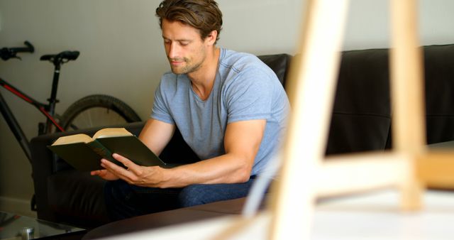 Young man reading book on living room couch. Casual attire and relaxed environment. Ideal for themes of learning, relaxation, solitude, and home life.