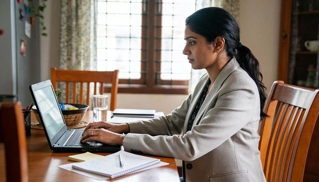Female professional working remotely at dining table, typing on laptop with notebook