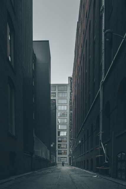 Solitary figure walking in narrow alley bordered by tall modern and older industrial buildings. Cold grey tones add to the sense of loneliness and drama. Ideal for city life themes, urban planning concepts, articles on solitude or the modern city's architectural contrast.